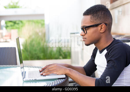 Closeup portrait, young nerdy man in big black glasses surfing the web on personal silver laptop, isolated outside outdoors back Stock Photo