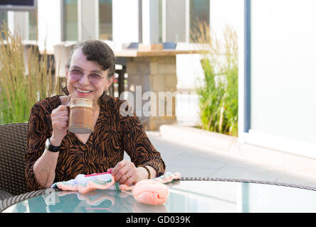 Closeup portrait, grandmother in brown dress, sitting, drinking, and crocheting, isolated outdoors outside background. Enjoying Stock Photo