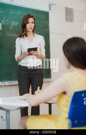 Smiling confident female teacher with a tablet-pc held in her hands standing in front of the blackboard Stock Photo