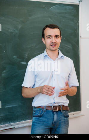 Portrait of young handsome student or teacher in a class pointing at blank chalkboard, talking and smiling Stock Photo