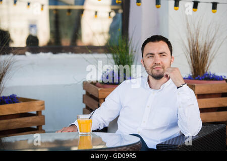 Handsome businessman is waiting for client in cafeteria. He is touching watch and looking to it with anticipation. The man is sitting at table outdoors and smiling. He is drinking orange juice Stock Photo