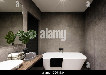 Modern bathroom wall made in dark color tiles which covered the bathtub and towels near the fancy plant Stock Photo