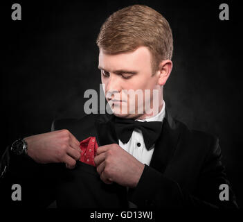 Young handsome man in suit with bow-tie adjusts his handkerchief on dark background. Stock Photo