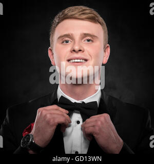Young handsome man in suit adjusts  bow-tie and smiling on dark background. Stock Photo