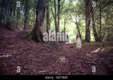 Woodland fantasy photography: a young red haired redhead woman girl wearing a long flowing dress walking down  a track away from an abandoned house by herself alone lost  in a forest woodland glade on a spring summer autumn afternoon, UK Stock Photo