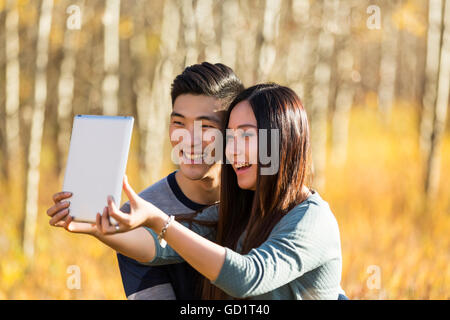 A Young Asian Couple Enjoying Quality Time Together Outdoors In A Park In Autumn And Taking Self-Portraits Using A Tablet In The Warmth Of The Sunl... Stock Photo