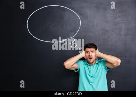 Shocked stressed young man with mouth opened covered ears by hands over chalkboard background Stock Photo