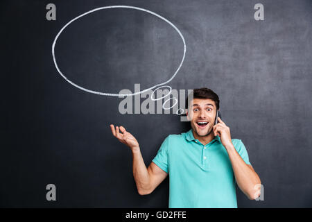 Surprised young man talking on mobile phone with thinking bubble on blackboard background Stock Photo