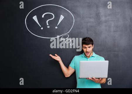 Surprised attractive young man holding and using laptop over blackboard background Stock Photo