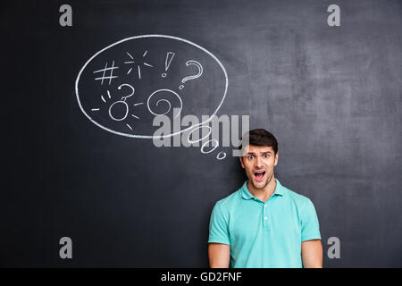 Confused shocked young man thinking about problem with blackboard behind him Stock Photo