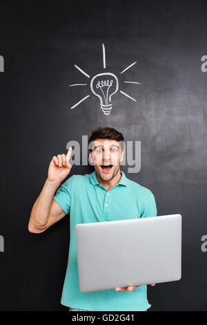 Excited young man pointing finger up and holding laptop over blackboard background with drawn light bulb Stock Photo