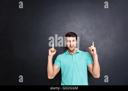 Smiling happy young man pointing up with both hands over black chalkboard Stock Photo