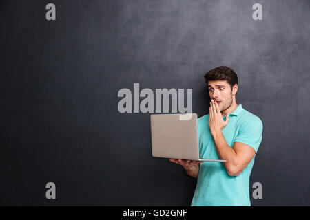 Amazed shocked young man standing and using laptop over chalkboard background Stock Photo