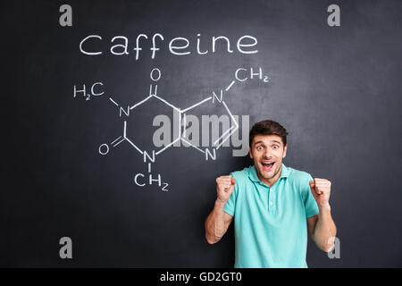 Happy excited young man celebrating success over drawn caffeine molecule chemical structure on chalkboard background Stock Photo