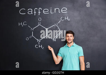 Smiling confident young student standing and showing chemical structure of caffeine molecule drawn on blackboard background Stock Photo