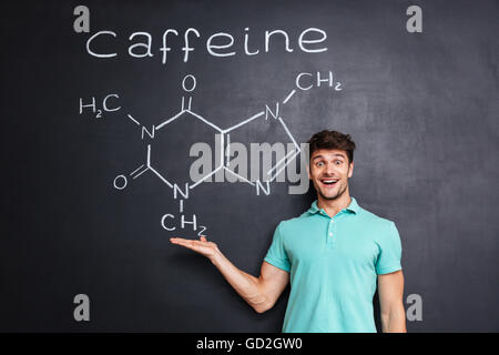 Cheerful young scientist showing chemical structure of caffeine molecule drawn on chalkboard background Stock Photo