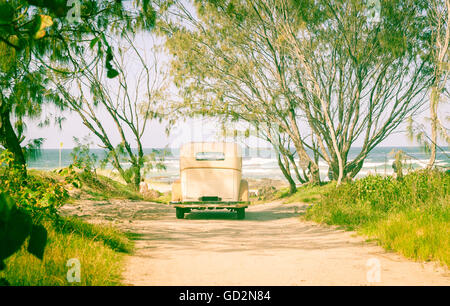 Vintage car parked at end of dirt track leading to beach under trees in retro filter tones. Stock Photo