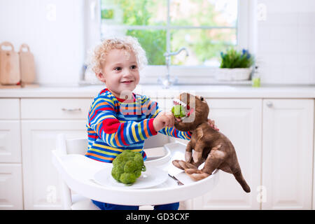 Happy little boy in high chair eating broccoli and feeding his toy dinosaur in a white kitchen. Stock Photo