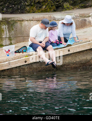 A family man woman and young child crab fishing on the slipway of the popular Cornish seaside town of Looe on a hot summers day wearing sun hats and looking into the harbour waters, Looe, Cornwall, England, UK Stock Photo