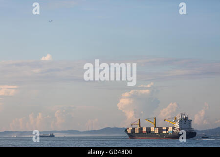 A container vessel armed with yellow cranes traveling out in the sea and a commercial air craft in sky. Singapore. Stock Photo