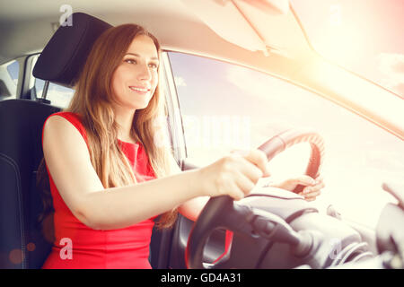 Young beautiful woman steering wheel driving a car. Confident smiling lady in red dress driving a car Stock Photo