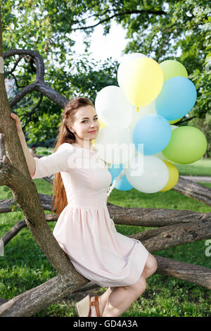 Beautiful young woman with balloons sitting on the tree in park. Happy caucasian girl in dress with bunch of bright balloons in  Stock Photo