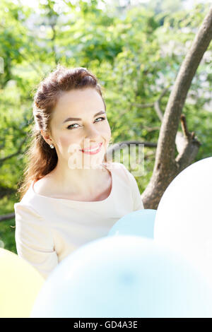 Beautiful young woman with balloons sitting on the tree in park. Happy caucasian girl in dress with bunch of bright balloons in  Stock Photo
