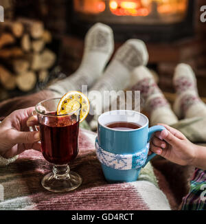 Warming and relaxing near fireplace. Woman feet near the cup of hot drink in front of fire. Stock Photo