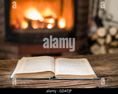 Opened book on the wooden table. Fireplace with warm fire on the background. Stock Photo