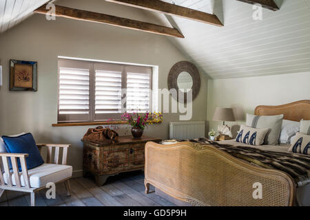 Plantation shutters and white wooden ceiling cladding in attic bedroom with vintage French cane bed. the chair is by Pomax Stock Photo