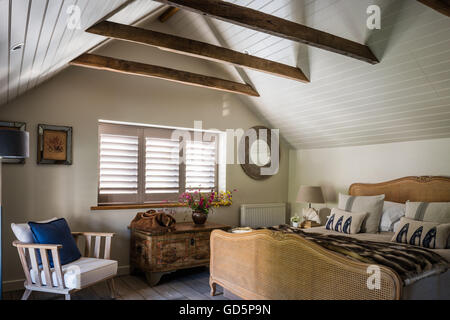 Plantation shutters and white wooden ceiling cladding in attic bedroom with vintage french cane bed. the chair is by Pomax Stock Photo