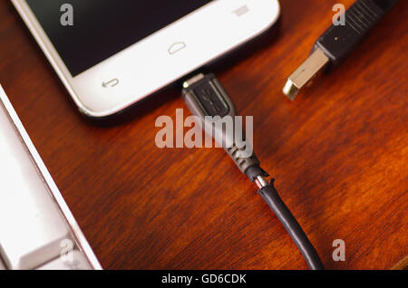 Broken usb mobile charger cable connected into smartphone lying on wooden surface next to computer keyboard, part of wire exposed through crack in plastic protection Stock Photo