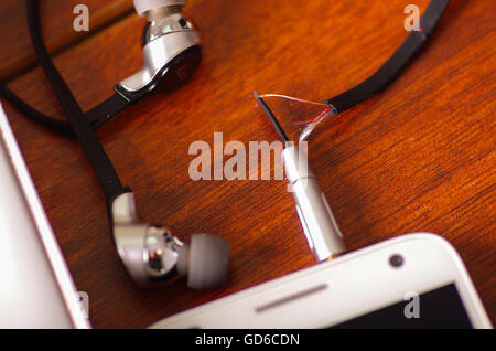 Broken audio headphones cable connected into smartphone lying on wooden surface, part of wire exposed through crack in plastic protection Stock Photo