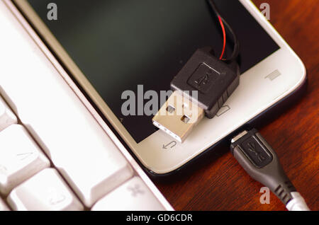 Broken usb mobile charger cable connected into smartphone lying on wooden surface next to computer keyboard, part of wire exposed through crack in plastic protection Stock Photo