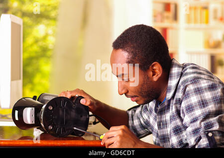 Man sitting by desk repairing small coffee maker using screwdiver, smiling happily while working Stock Photo