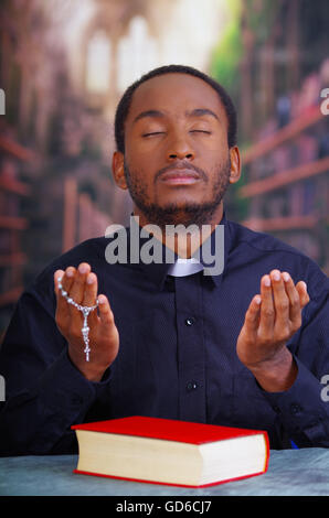 Catholic priest wearing traditional clerical collar shirt sitting with folded hands holding rosary while praying, book on desk in front, religion concept Stock Photo