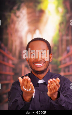 Catholic priest wearing traditional clerical collar shirt standing facing camera, holding hands out with rosary cross, looking forward, religion concept Stock Photo
