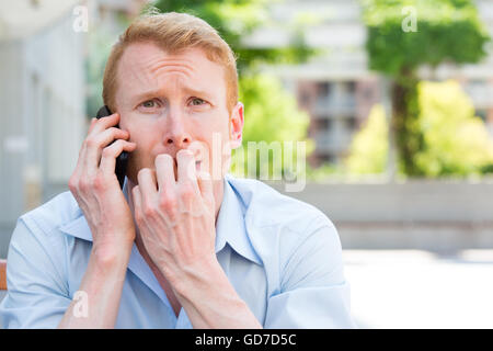 Closeup portrait, young man biting finger nails, worried about something he hears on phone, isolated outdoors background Stock Photo