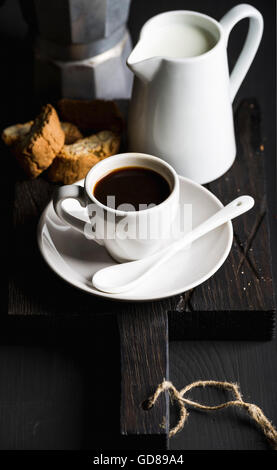 Italian coffee breakfat set. Cup of hot espresso, creamer with milk and cookies on dark rustic wooden board over black backgroun Stock Photo