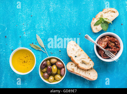 Mediterranean snacks set. Pickled olives, oil, sun-dried tomatoes, herbs and sliced ciabatta bread over blue painted background, Stock Photo
