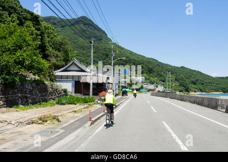 Two touring cyclists riding on a country road past a traditional Japanese village on Shodo Island. Stock Photo