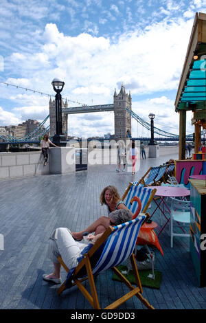Londoners relax in deck chairs at a restaurant situated on the walkway along the Thames Tower Bridge a London landmark in the background Stock Photo
