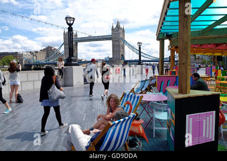 Londoners relax in deck chairs at a restaurant situated on the walkway along the Thames Tower Bridge a London landmark in the background Stock Photo