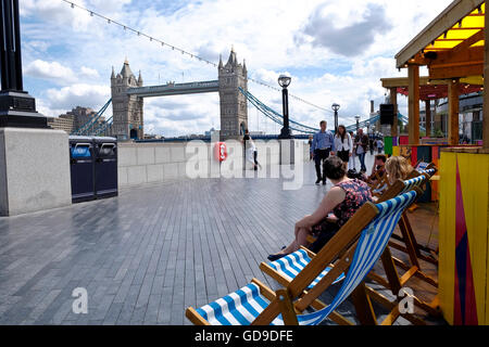 Tourists sit and relax in deck chairs on the walkway on the south bank of the Thames river with Tower Bridge a London landmark in the background. Stock Photo
