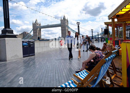 London, United Kingdom. Tourists sit and relax in deck chairs on the walkway on the south bank of the Thames Tower Bridge a London landmark Stock Photo