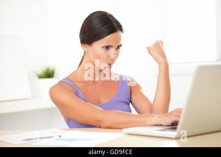 Excited young woman in purple shirt sitting and working on laptop as freelancer at home Stock Photo