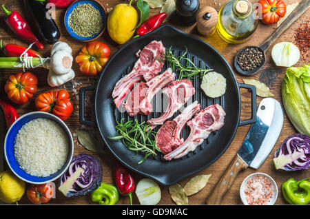 Ingredients for cooking healthy meat dinner. Raw uncooked lamb chops in dark grilling iron pan, vegetables, rice, herbs, olive o Stock Photo