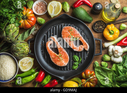 Raw uncooked salmon steakes with different vegetables, rice, herbs and spices on black grilling iron pan over rustic wooden back Stock Photo