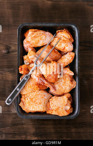 Raw Marinated chicken meat wings and legs for BBQ, served in plastic box with vintage meat fork over dark wooden background. Top Stock Photo