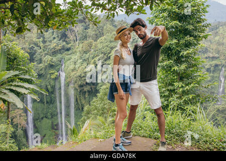 Beautiful young couple standing together on a cliff and taking selfie with waterfall. Man and woman in forest taking self portra Stock Photo